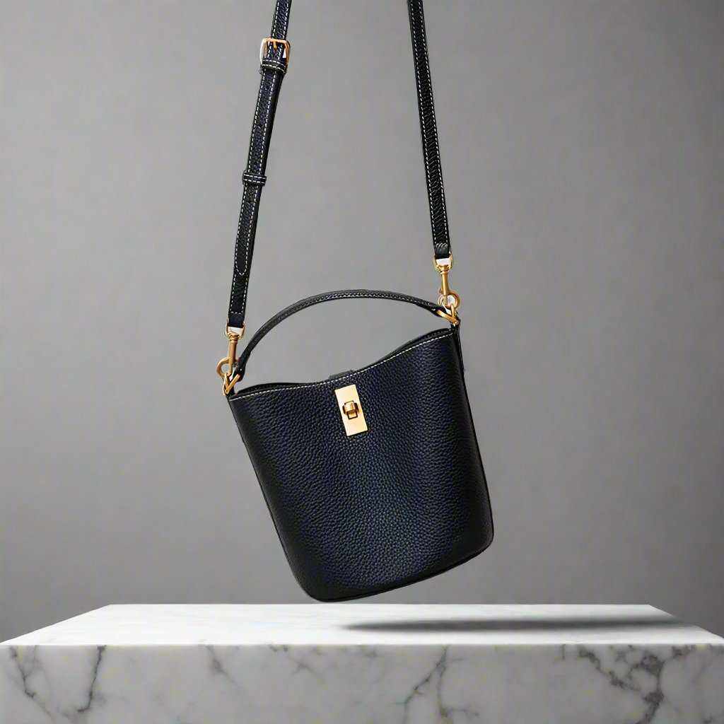 Black leather crossbody bucket purse with adjustable strap and gold hardware, displayed on a marble surface.