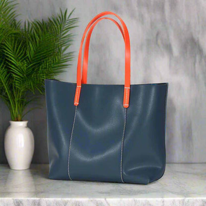 Blue leather handbag with contrasting orange handles on a marble surface, featuring green plant and gray background.