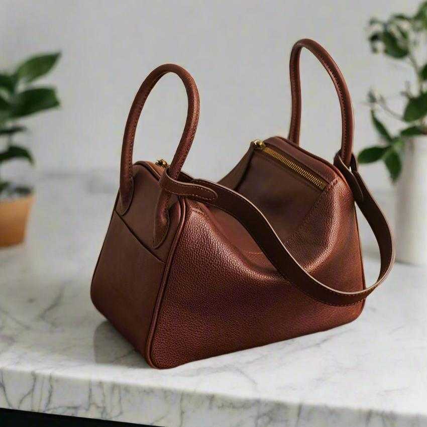 Brown leather satchel purse with dual handles and a zip closure, displayed on a marble surface.