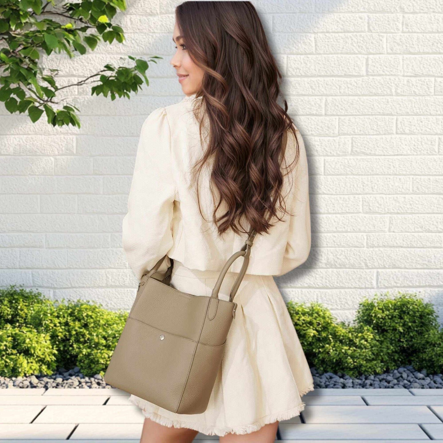 Woman wearing a white dress and carrying a leather tote bag crossbody in light brown, featuring a bucket-style design, outdoors against a white brick wall.