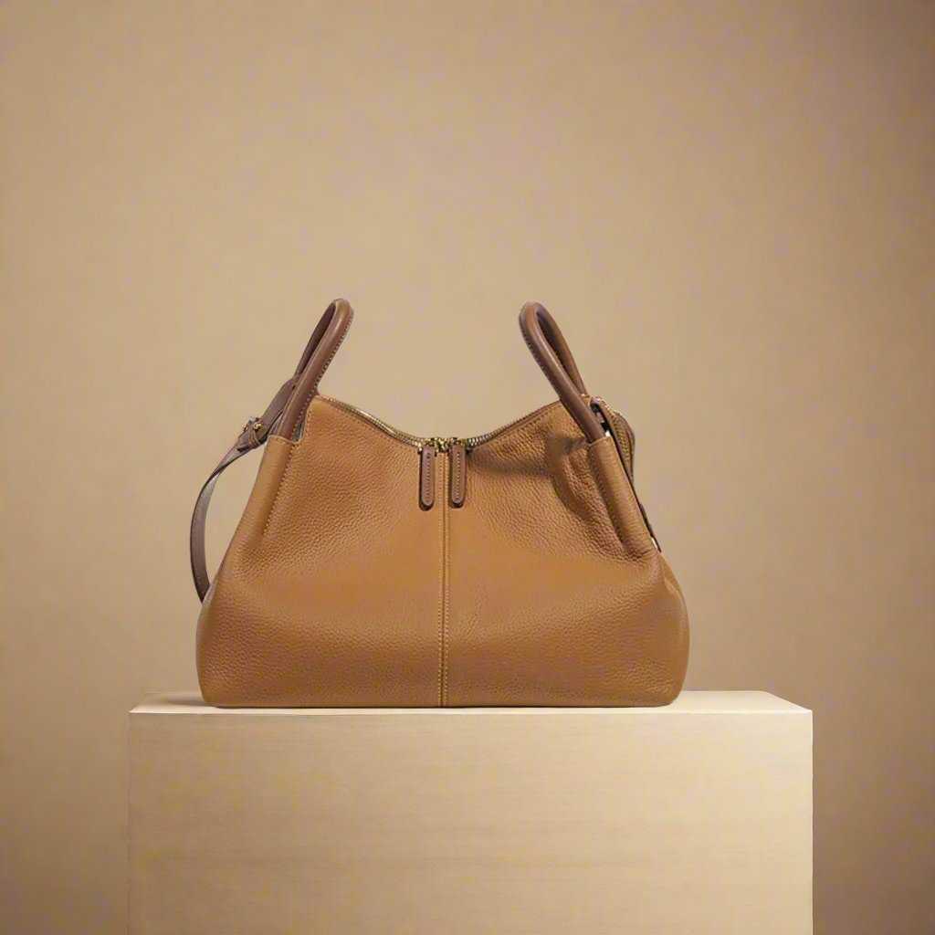 Soft leather crossbody bag with dual handles in brown, displayed on a minimalist stand.