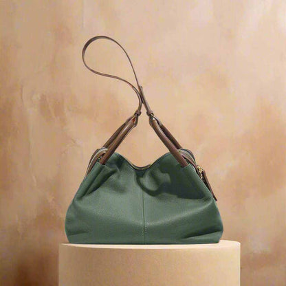 Soft leather crossbody bag in green with dual handles and removable shoulder strap, displayed on pedestal.