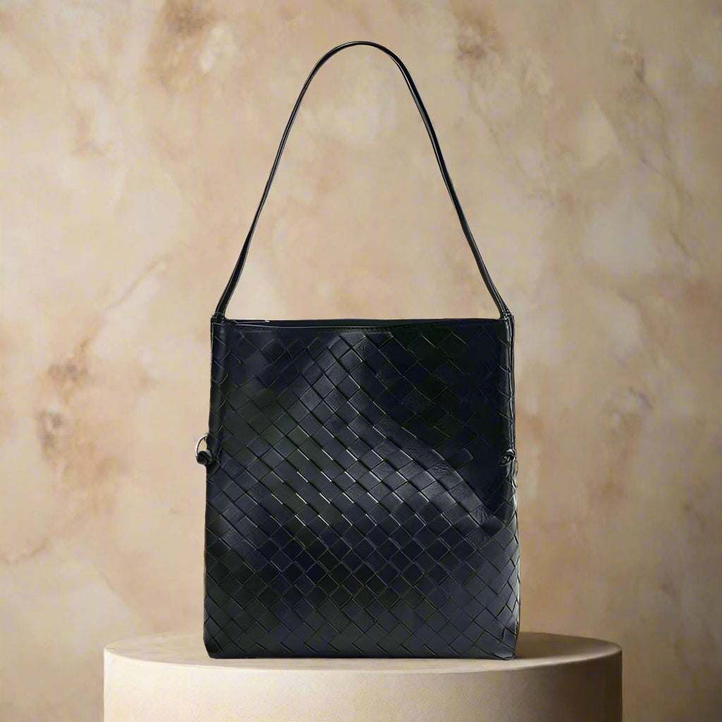 Woven leather tote handbag in dark color displayed on a pedestal.